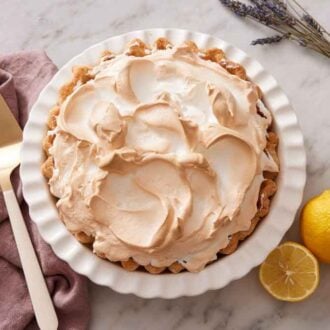 A slightly overhead view of a lemon meringue pie in a white baking dish. Lemon, lavender, and a cake spatula off on the side.