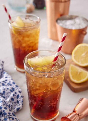 Two glasses of Long Island Iced Tea with sliced lemon and straws. Lemon lemons and ice in the background with a shaker and jigger.