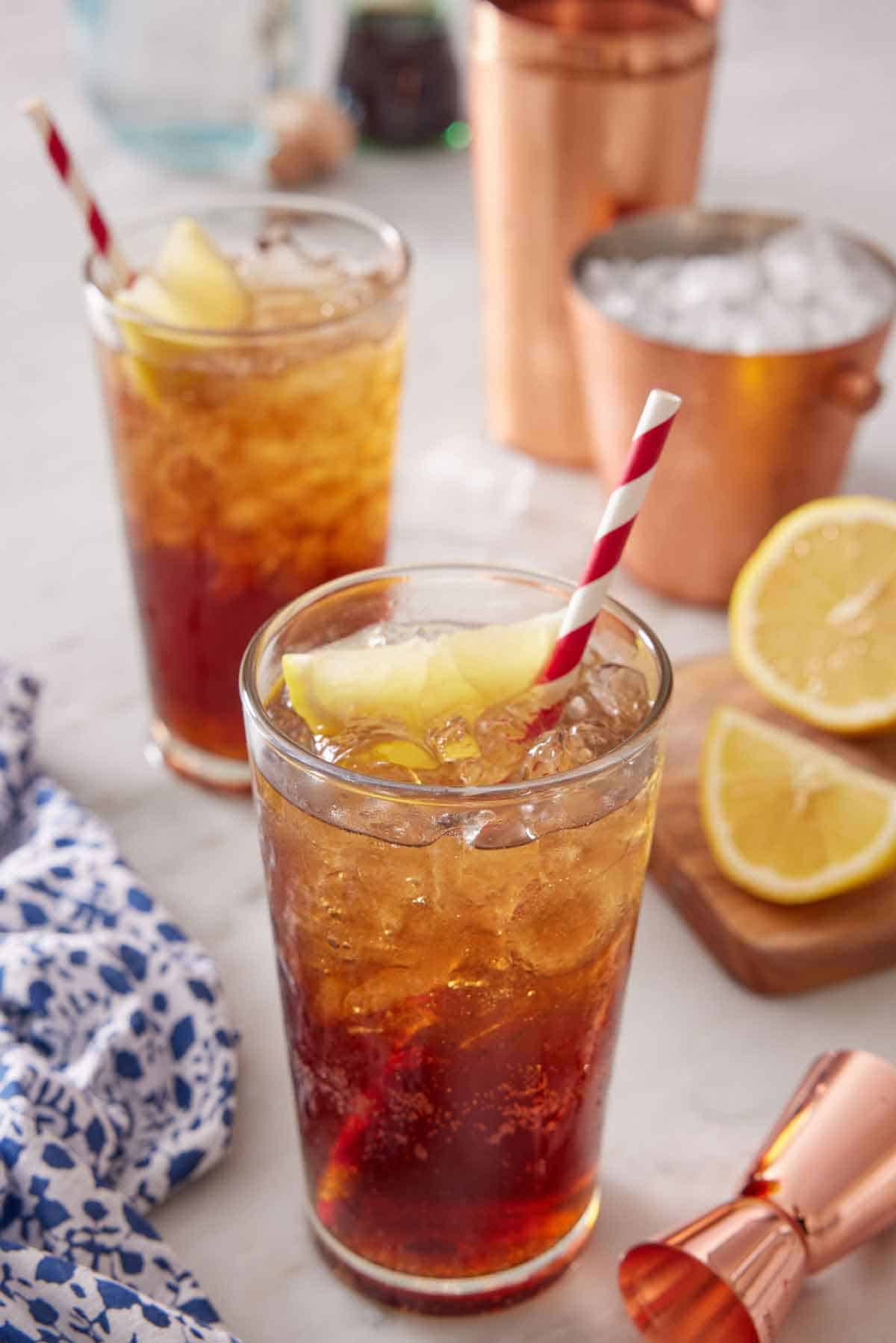 Two glasses of Long Island Iced Tea with sliced lemon and straws. Lemon lemons and ice in the background with a shaker and jigger.