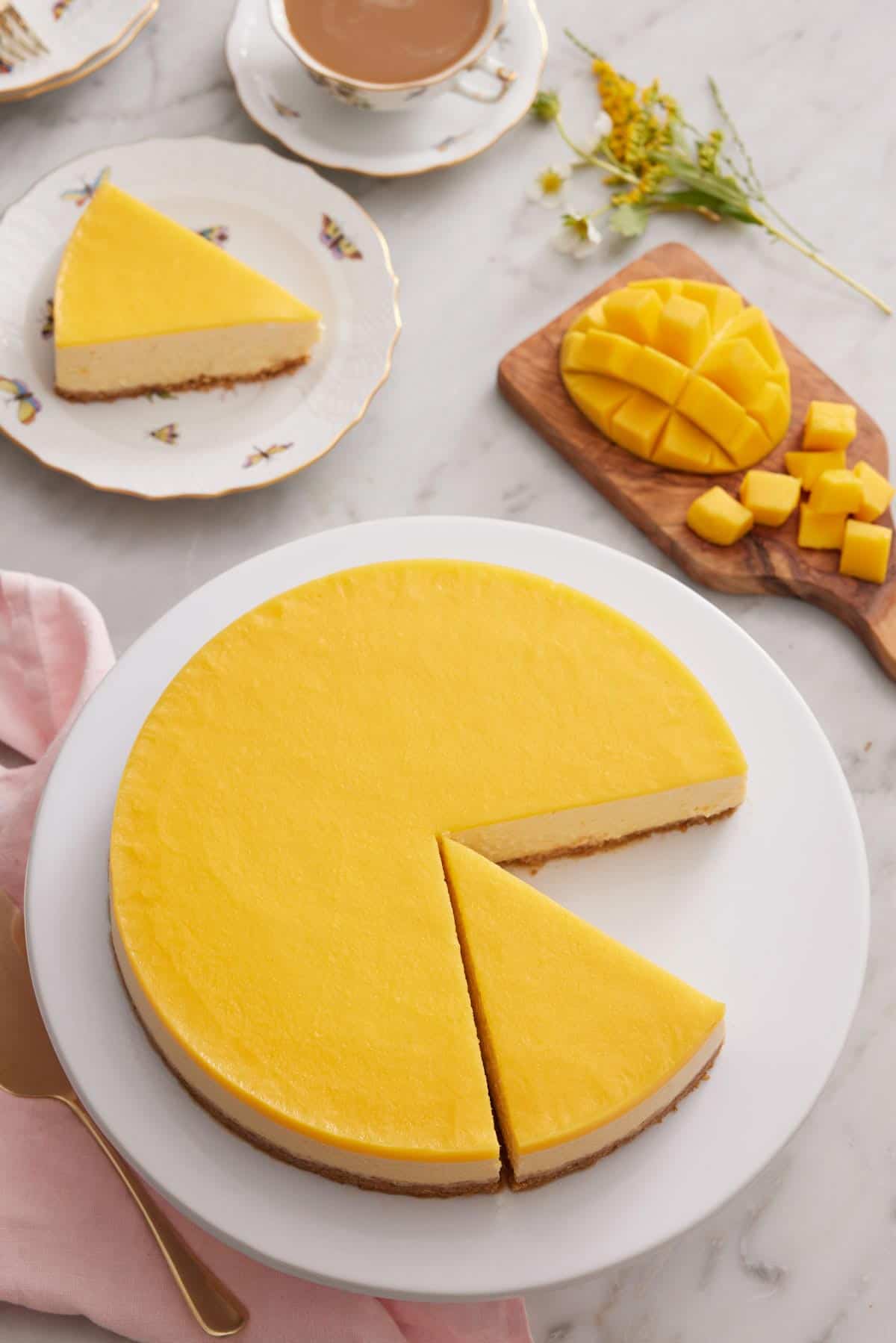 Overhead view of a mango cheesecake with one slice removed and one sliced cut. Plated slice in the background along with a diced mango.
