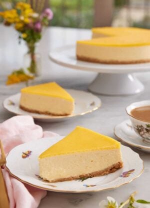 Two slices of mango cheesecake on two plates with a cake stand in the background with the rest of the cake.