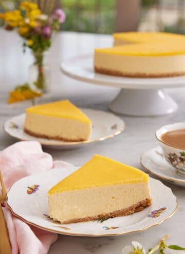 Two slices of mango cheesecake on two plates with a cake stand in the background with the rest of the cake.