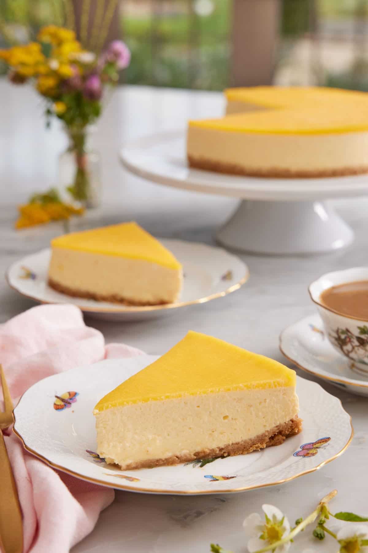 Two slices of mango cheesecake on two plates with a cake stand in the background with the rest of the cake.