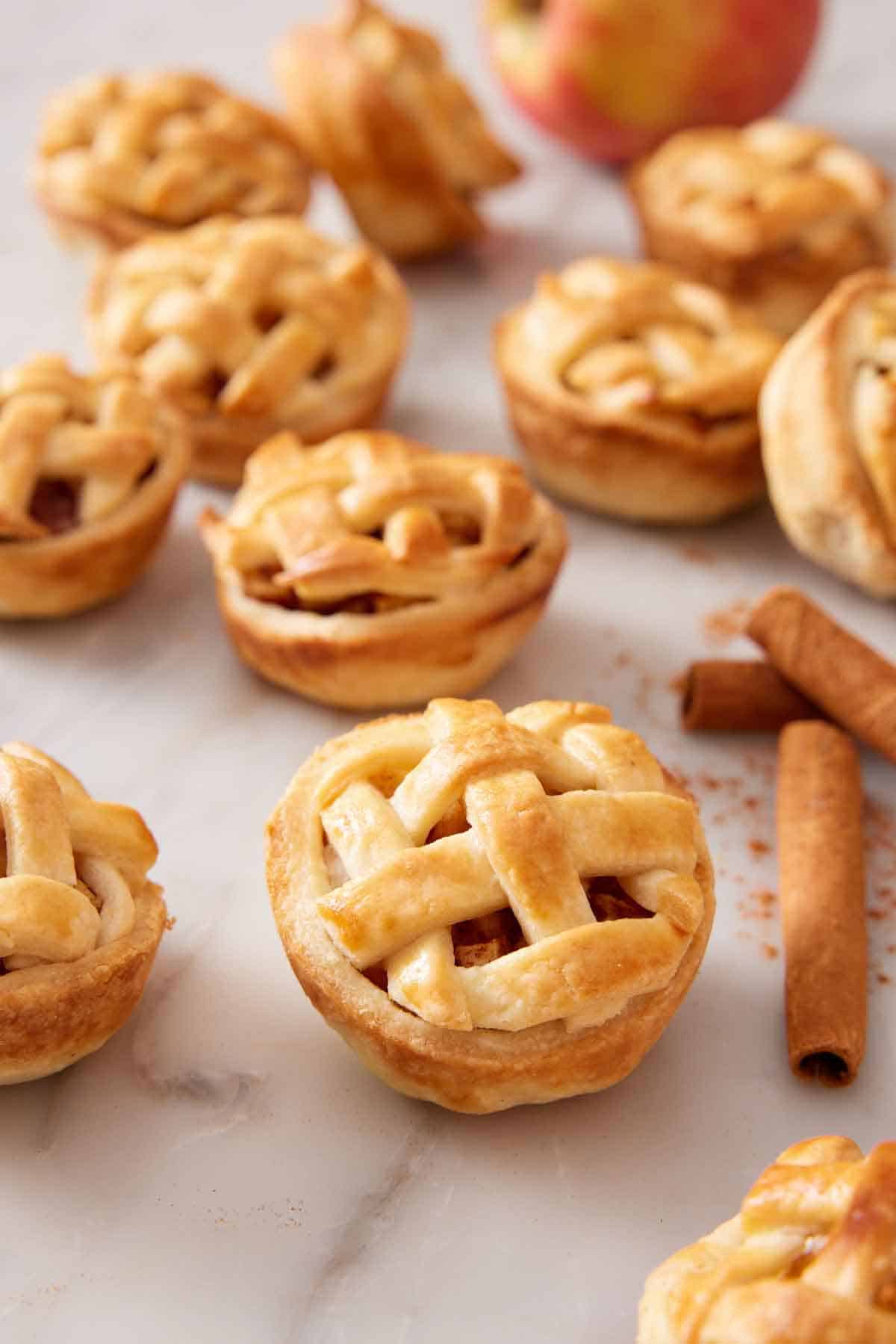 Multiple mini apple pies on a marble surface with cinnamon sticks on the side.