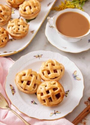 A plate with three mini apple pies with a cup of coffee and platter or more mini apple pies behind it.