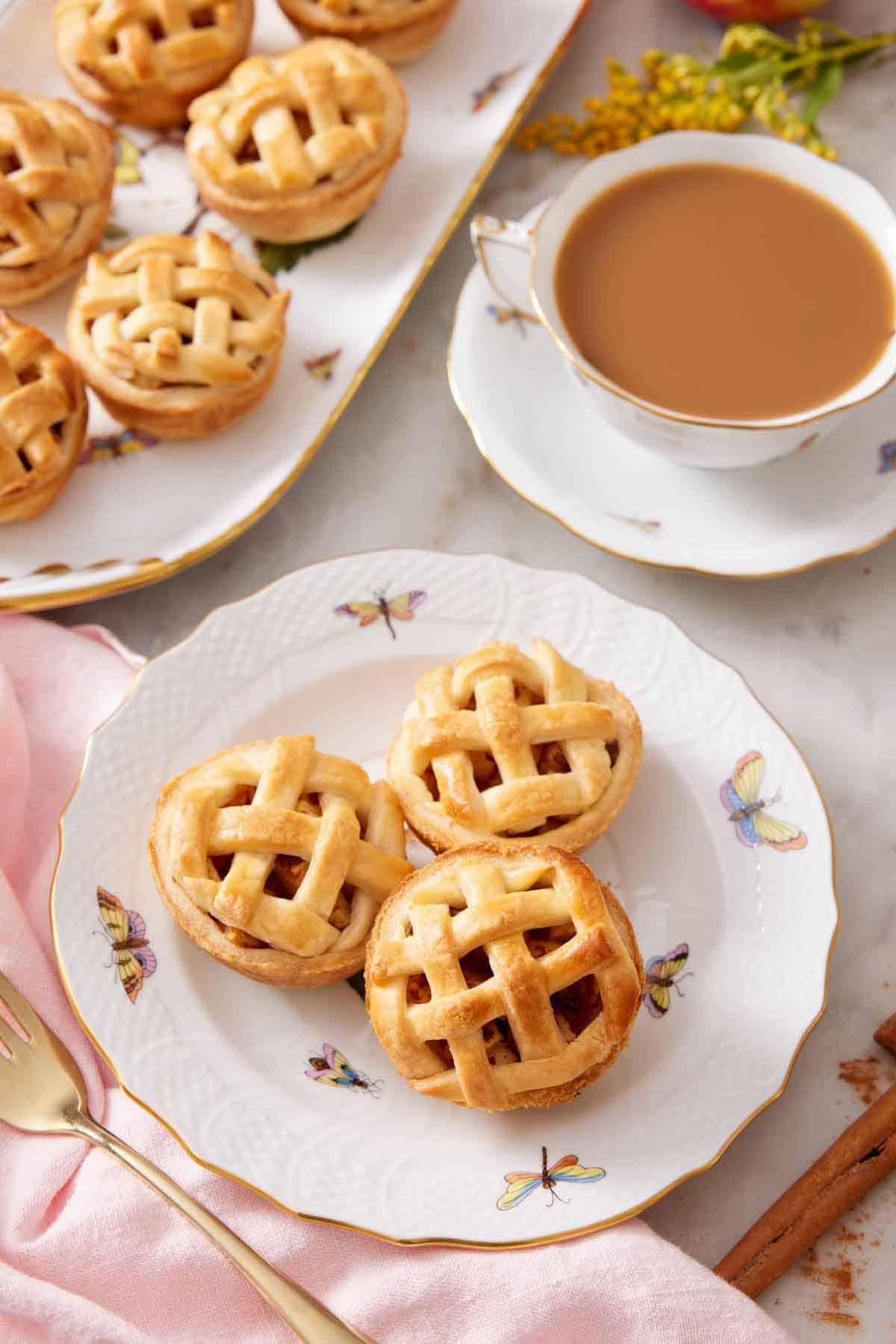 A plate with three mini apple pies with a cup of coffee and platter or more mini apple pies behind it.