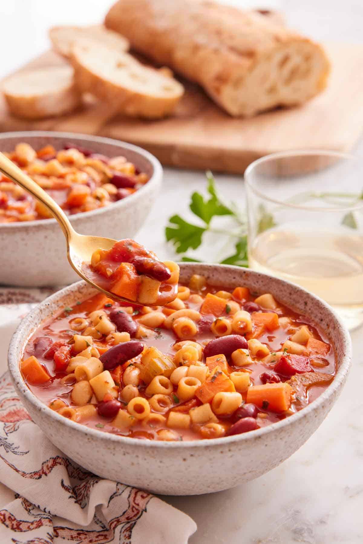 A bowl of pasta fagioli with a spoonful lifted out with wine and bread in the background.