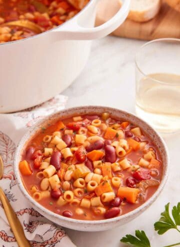 A bowl of pasta fagioli with a pot and glass of wine in the background.