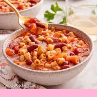 Pinterest graphic of a bowl of pasta fagioli with a spoonful lifted out with wine and bread in the background.