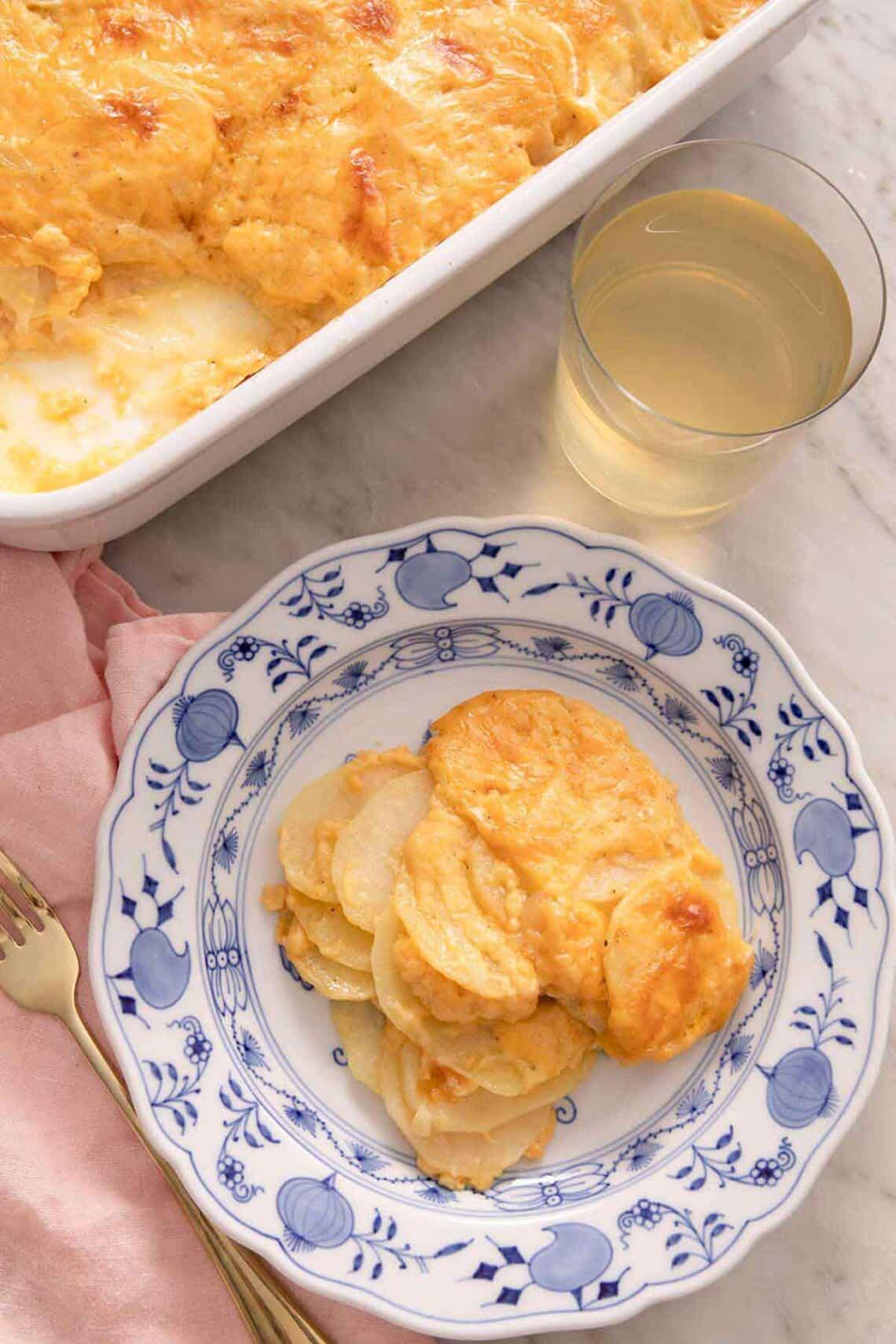 An overhead view of a plate of potatoes au gratin with a glass of wine being it along with a casserole dish.