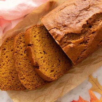 A loaf of pumpkin bread with three slices cut over brown parchment paper and a pink napkin.