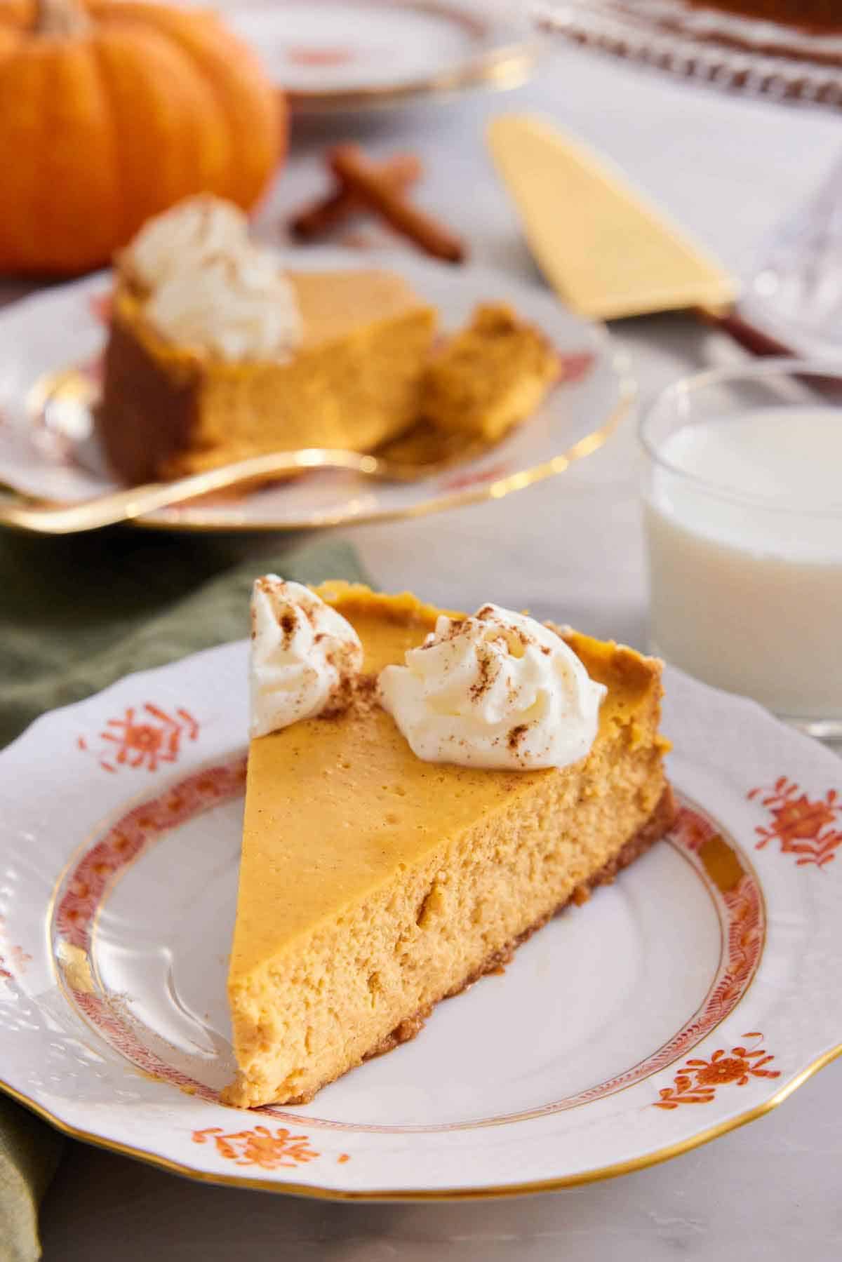 A slice of pumpkin cheesecake on a plate topped with whipped cream. A glass of milk and second slice in the background.