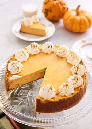 A pumpkin cheesecake on a cake stand with a slice removed. Mini pumpkins and a plated slice in the background.