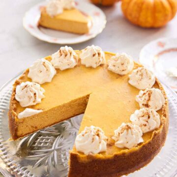A pumpkin cheesecake on a cake stand with a slice removed. Mini pumpkins and a plated slice in the background.