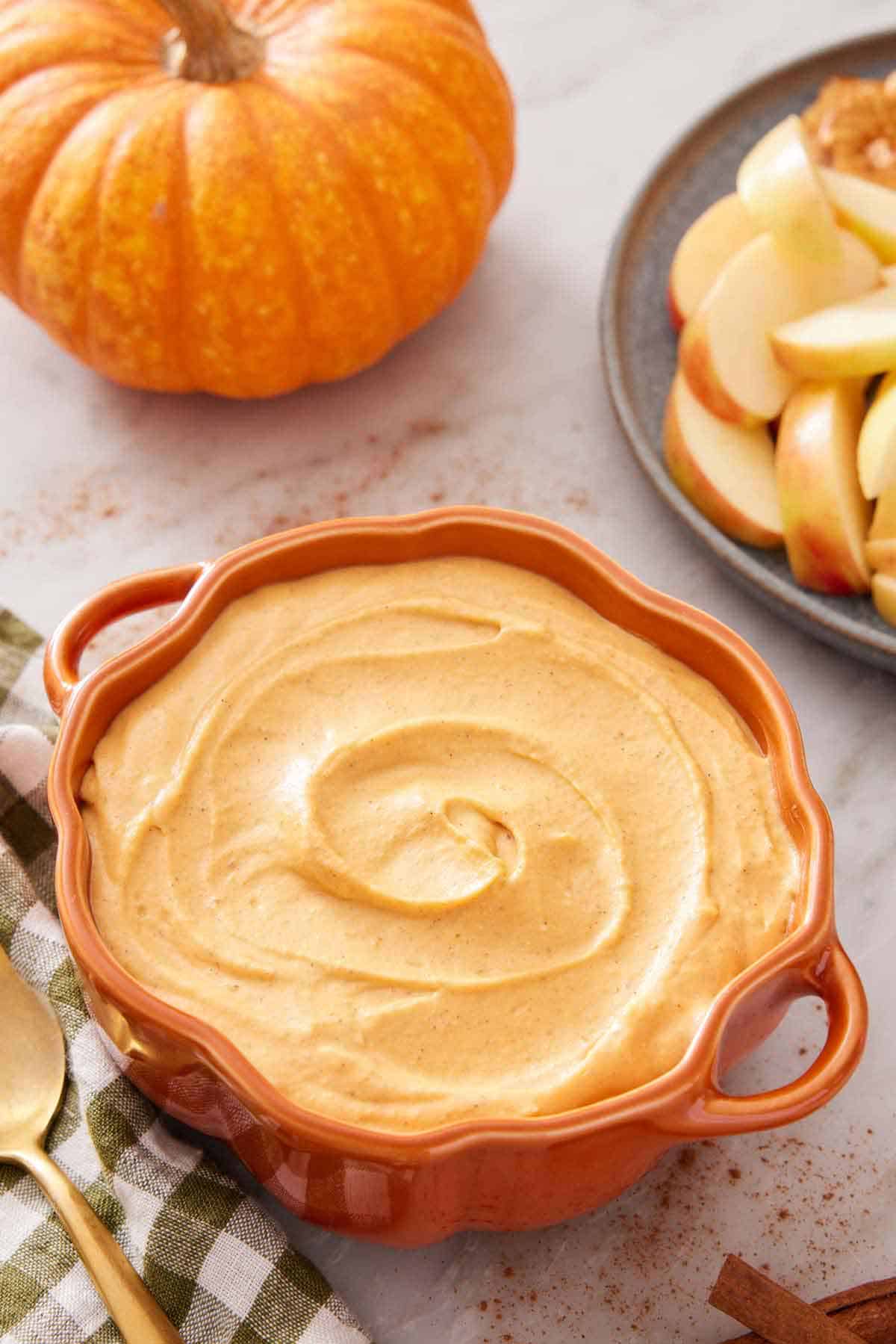 A pumpkin-shaped bowl containing pumpkin dip with some apple slices and a pumpkin on the side.