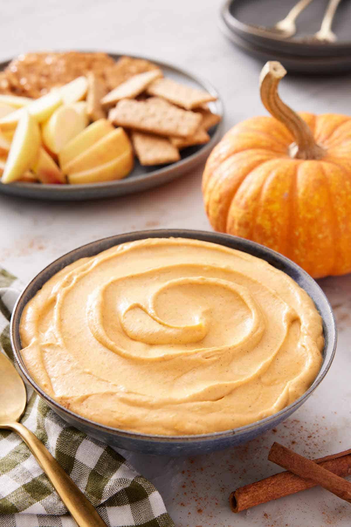 A bowl of pumpkin dip with crackers, apples, and pumpkin in the background.