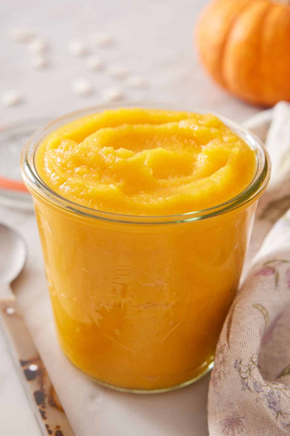 A glass jar of pumpkin puree with a spoon on the side and a pumpkin in the background.