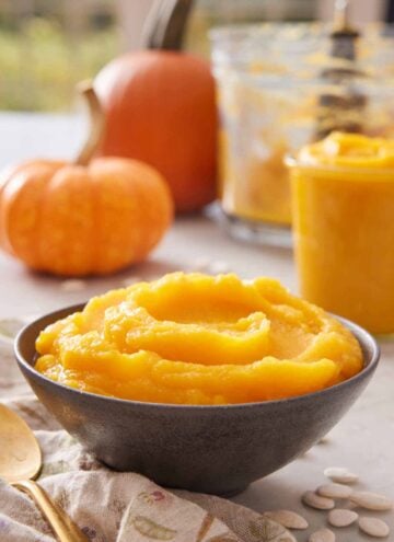A bowl of pumpkin puree with some pumpkins in the background alongside a food processor bowl and a jar of puree.