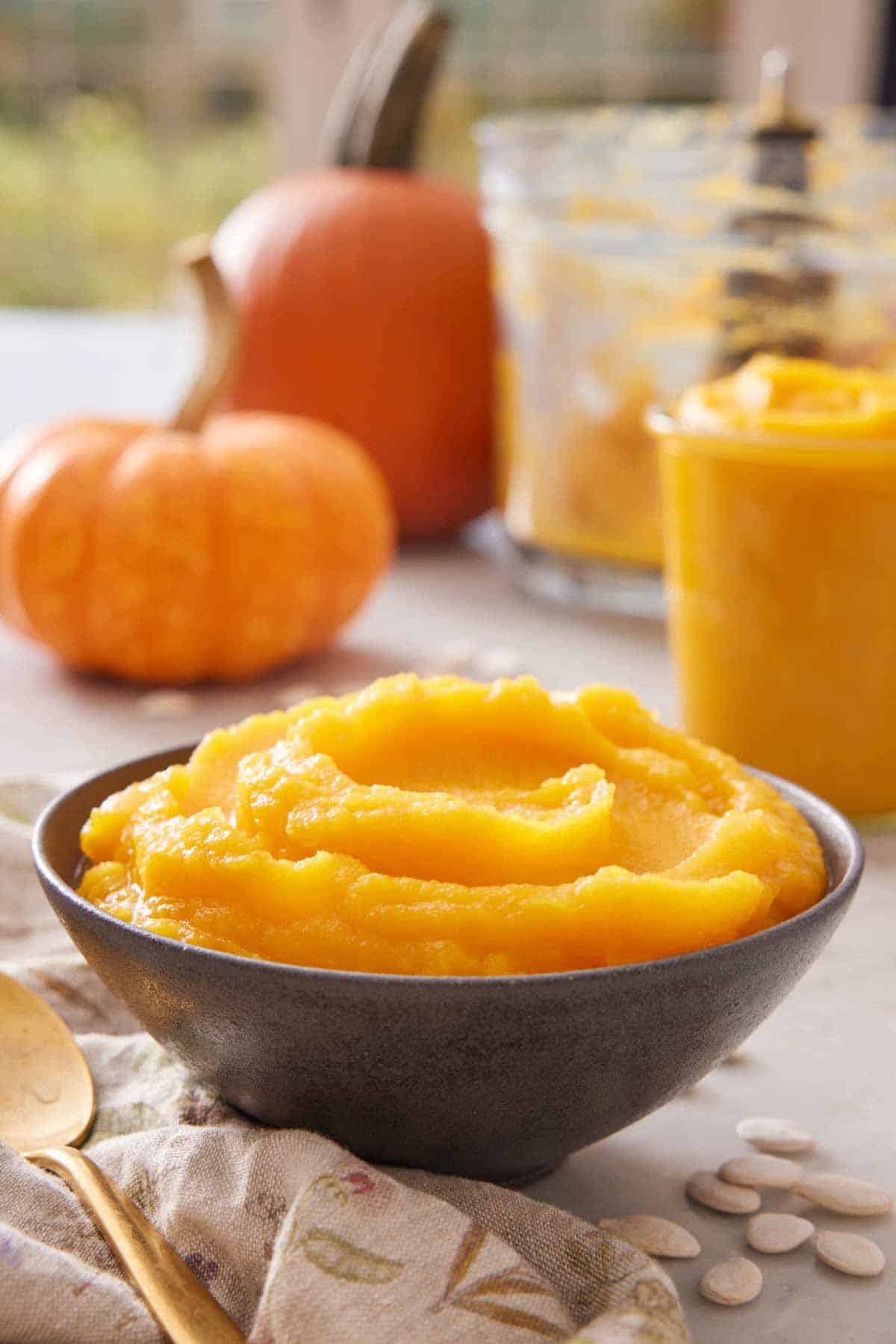 A bowl of pumpkin puree with some pumpkins in the background alongside a food processor bowl and a jar of puree.