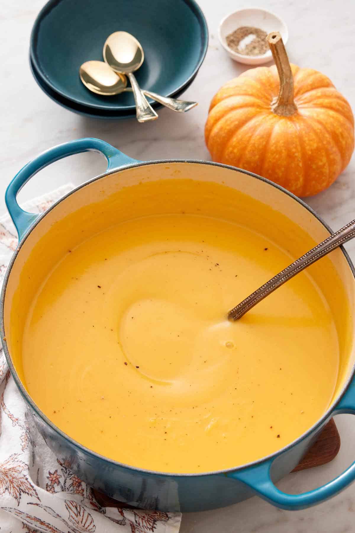 A large pot of pumpkin soup with a ladle inside. Some bowls, spoons, and a pumpkin in the background.