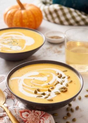 Two bowls of pumpkin soup with a drizzle of cream and pumpkin seeds as a garnish and a glass of wine in the background along with a pumpkin.