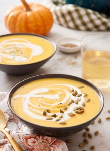 Two bowls of pumpkin soup with a drizzle of cream and pumpkin seeds as a garnish and a glass of wine in the background along with a pumpkin.