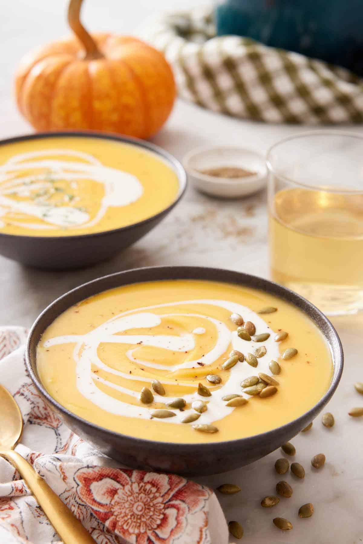 Two bowls of pumpkin soup with a drizzle of cream and pumpkin seeds as a garnish and a glass of wine in the background along with a pumpkin.