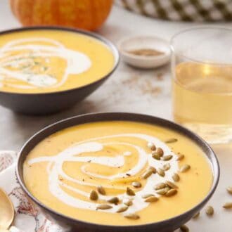 Pinterest graphic of two bowls of pumpkin soup with a drizzle of cream and pumpkin seeds as a garnish and a glass of wine in the background along with a pumpkin.