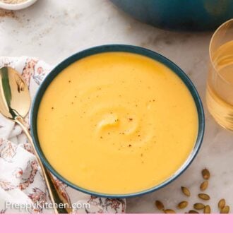 Pinterest graphic of a bowl of pumpkin soup topped with some pepper. Wine, pot, bowl of pepper, and spoon off to the side.