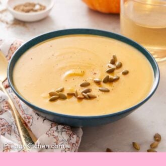 Pinterest graphic of a bowl of pumpkin soup with some pumpkin seeds as garnish with a glass of wine and a pumpkin in the background.