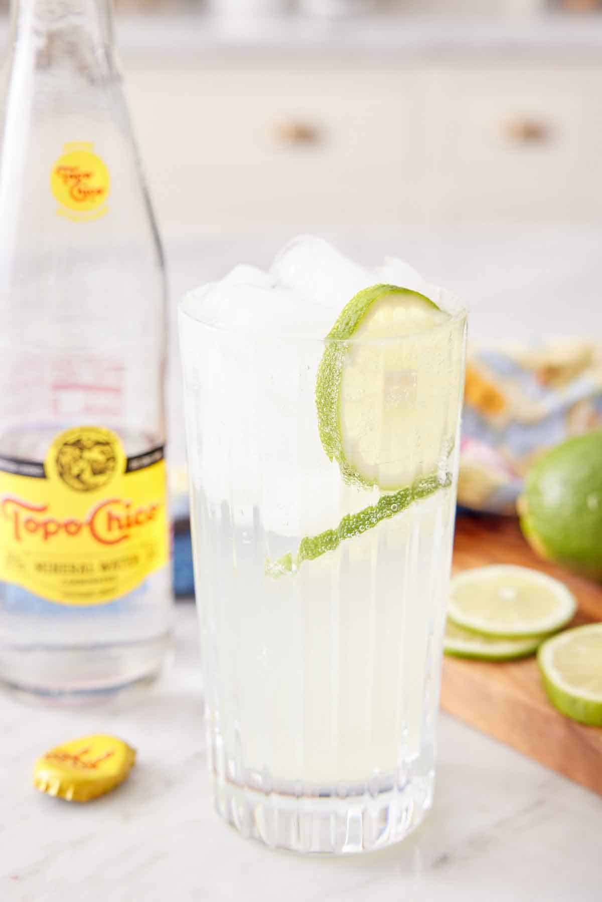 A glass of ranch water with lime slices in the drink with a glass of topo chico in the background.