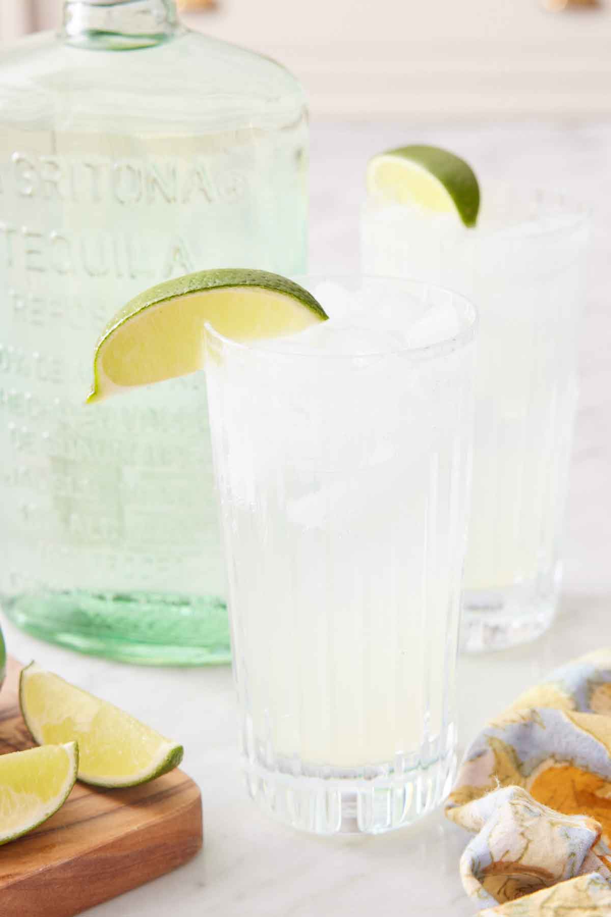 Two glasses of ranch water with lime wedges on the rim and a bottle of tequila in the back.