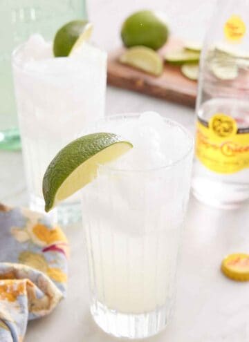Two glasses of ranch water with lime wedges on the rim. A glass of topo chico in the background along with cut limes.