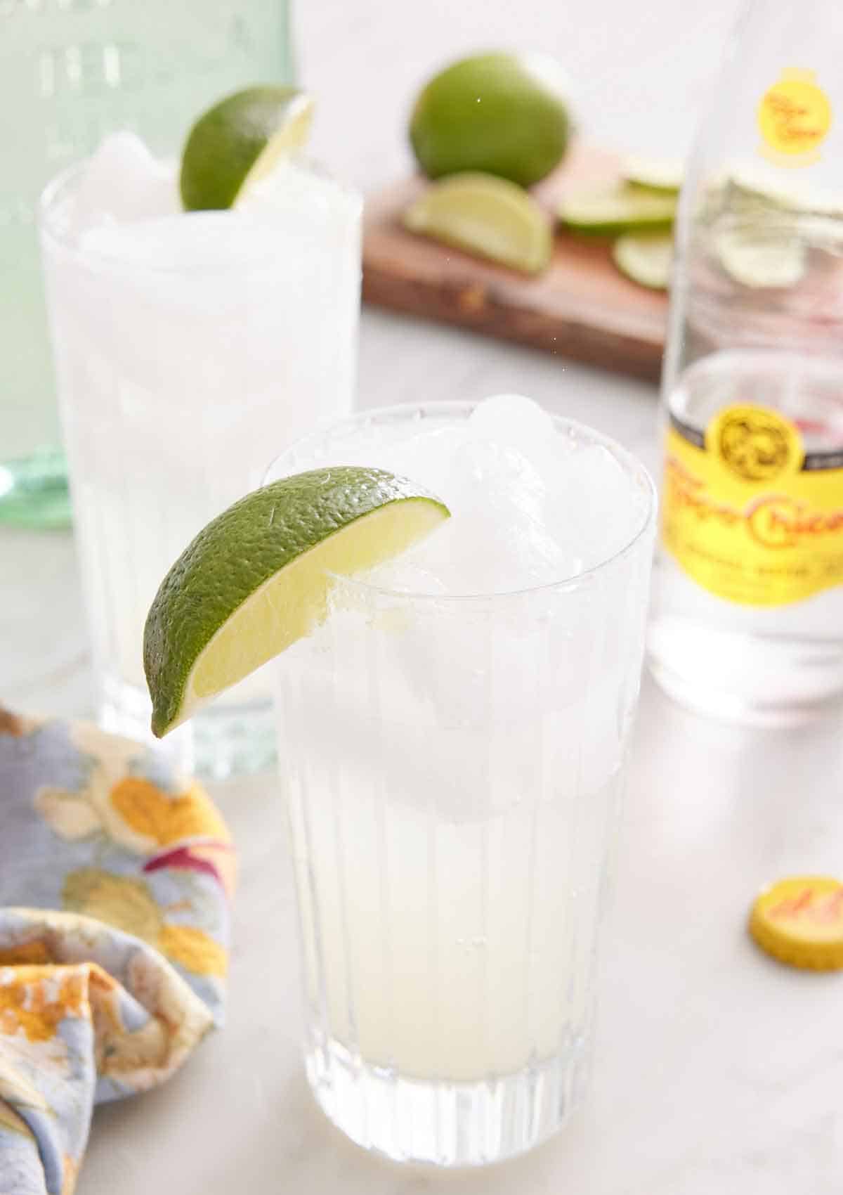 Two glasses of ranch water with lime wedges on the rim. A glass of topo chico in the background along with cut limes.