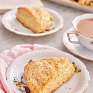 A plate with a scone with a mug of coffee, another plate with a scone, a platter of scones, and an opened book in the background.