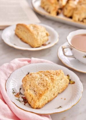 A plate with a scone with a mug of coffee, another plate with a scone, a platter of scones, and an opened book in the background.