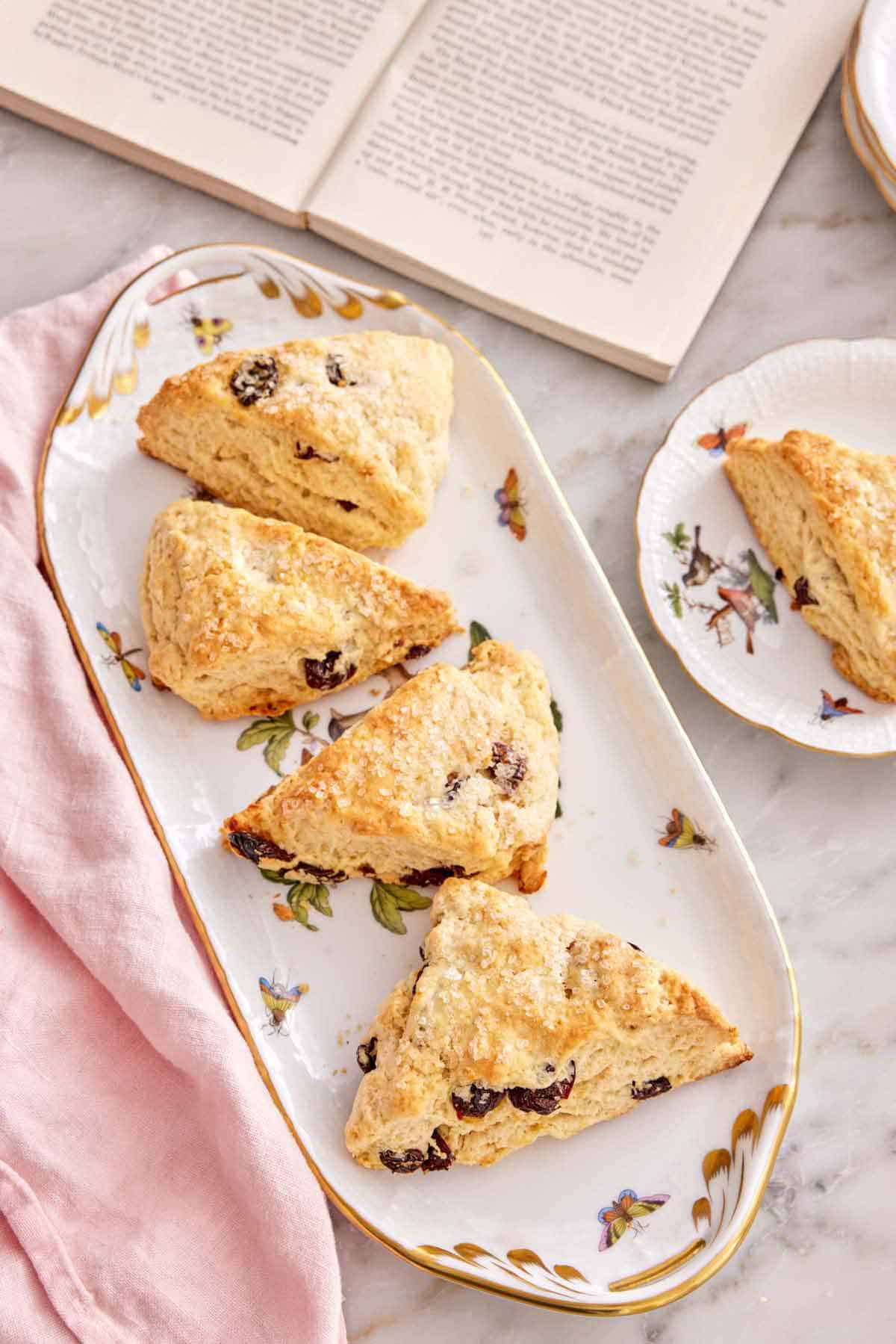 Overhead view of a platter with four scones by an opened book and a scone on a plate.