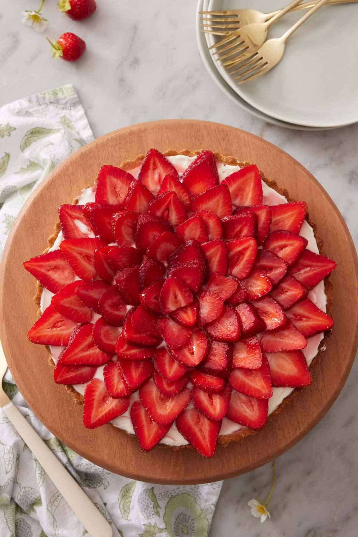 Overhead view of a strawberry tart with a stack of plates and forks to the side.