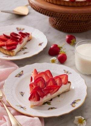 A plate with a slice of strawberry tart with strawberries, glass of milk, a second plated slice, and cake stand in the background.