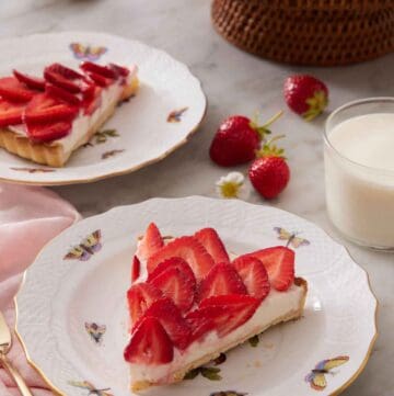 A plate with a slice of strawberry tart with strawberries, glass of milk, a second plated slice, and cake stand in the background.