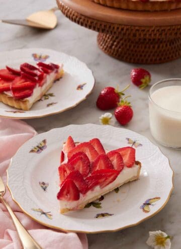 A plate with a slice of strawberry tart with strawberries, glass of milk, a second plated slice, and cake stand in the background.