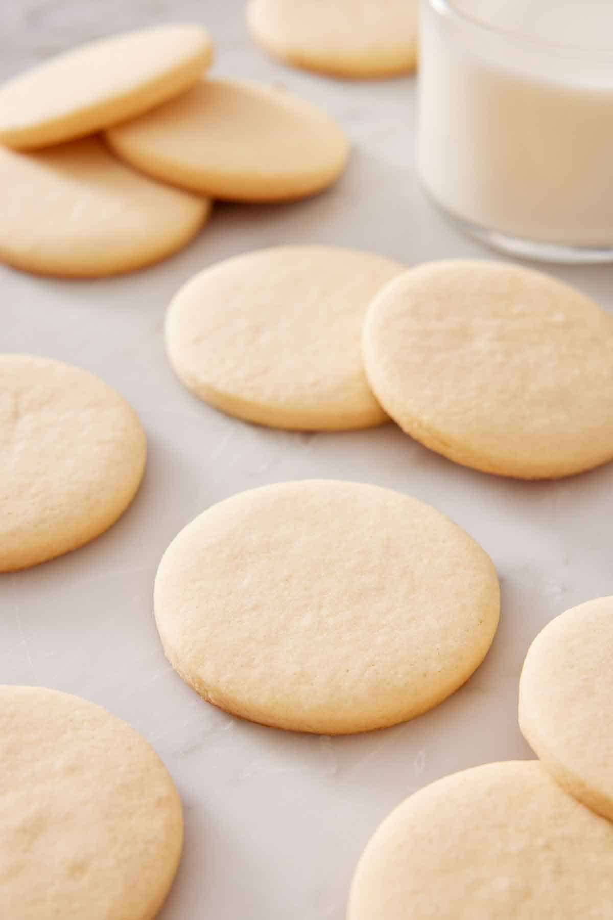 Multiple sugar cookies on a marble surface, some are slightly stacked on others. A glass of milk in the background.