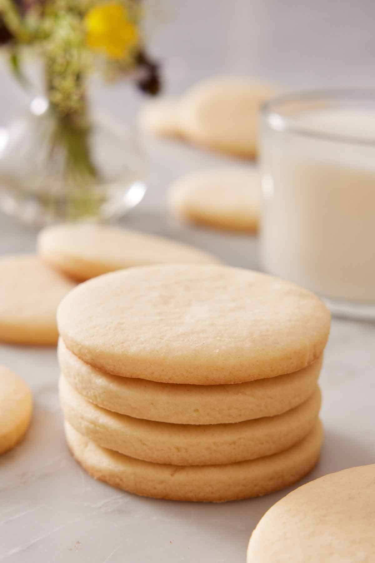 A stack of four sugar cookies with a glass of milk in the background and more cookies scattered around.