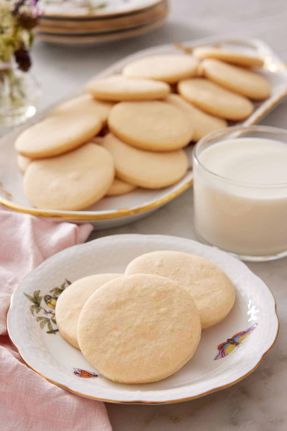 A plate with three sugar cookies with a glass of milk and additional cookies on a platter in the background.