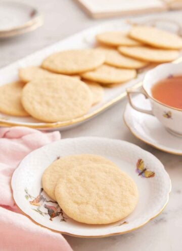A plate with two tea cakes with a mug of tea and platter of more cookies in the back.