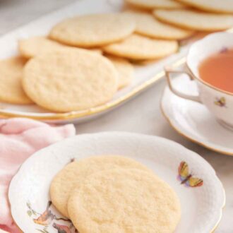 Pinterest graphic of a plate with two tea cakes with a mug of tea and platter of more cookies in the back.