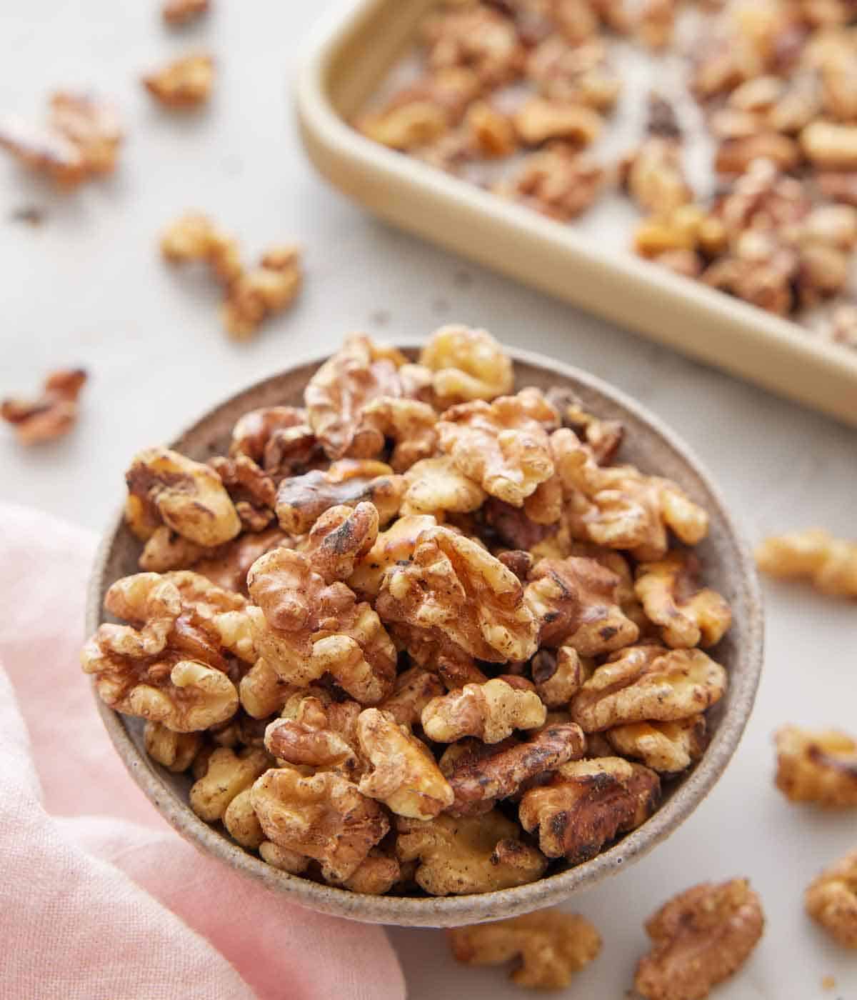 A bowl of toasted walnuts with a sheet pan in the background with more walnuts scattered.