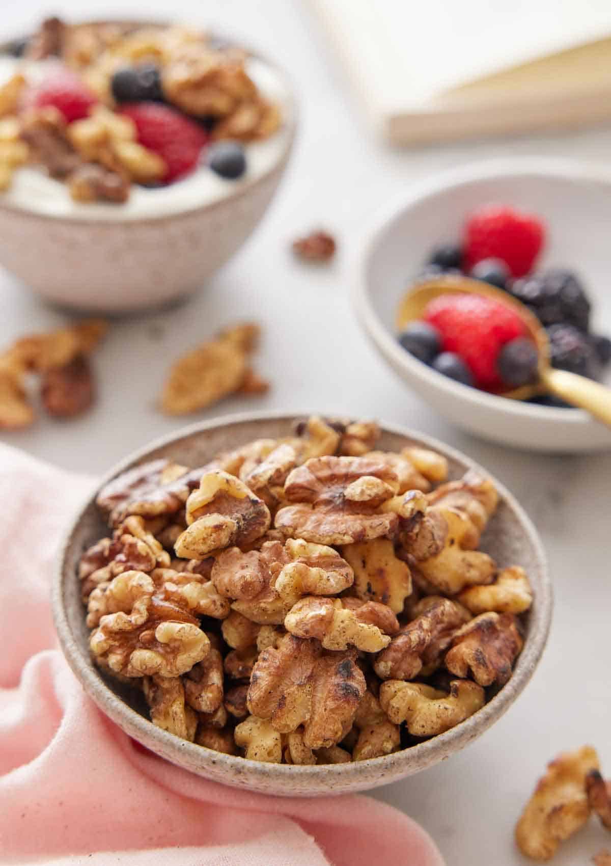 A bowl of toasted walnuts with some berries in the background along with a bowl of yogurt with toasted walnuts and berries.