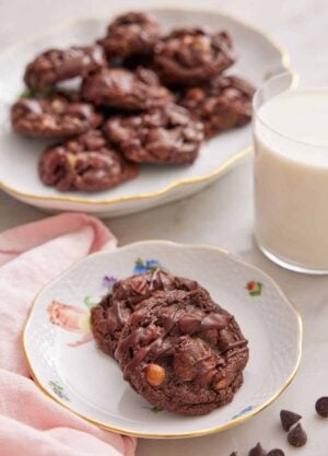 A plate with two turtle cookies with a glass of milk and platter of cookies in the background.
