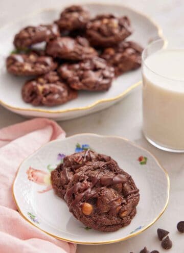A plate with two turtle cookies with a glass of milk and platter of cookies in the background.